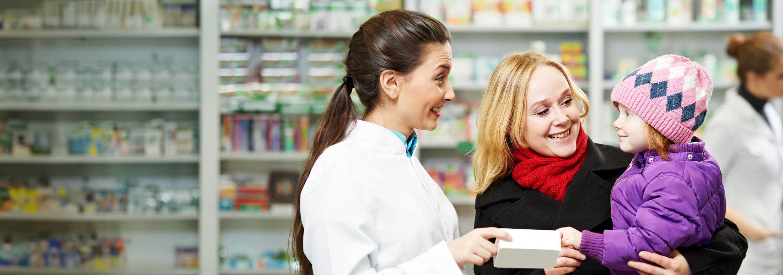 Eine Apothekerin hilft einer Frau in Rot mit einem Kind, das eine rosa Mütze trägt, in einer Apotheke, die von Regalen mit Gesundheitsprodukten umgeben ist.